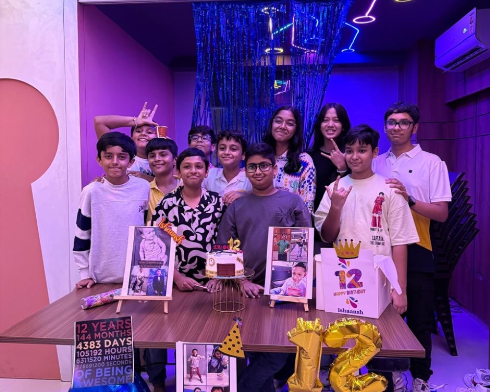 A group of kids posing together at Escape Zone during a 12th birthday celebration, standing behind a decorated table with cake, balloons, and birthday props in a vibrant, neon-lit party room.
